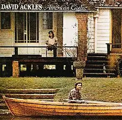 A photo of a man in a long, wooden canoe, in front of a house with a woman sat on the porch railing.