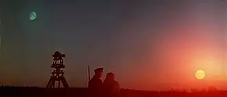 Two men stand between the sun and the moon with one rising as the other sets
