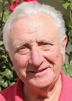 Colour press shot of a clean-shaven, smiling man with short, white hair, combed back