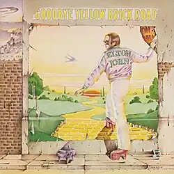 A wall with a worn poster showing a yellow brick road winding down towards the green plains and the sunset. Elton John, shown with a purple jacket bearing his name, has his right foot on the yellow brick road and his right hand holding on to the poster. On the pavement, a miniature piano with a musical note. The album's title is placed above the poster.