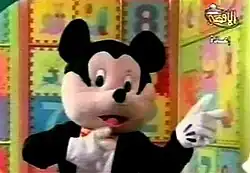 A giant costumed character with a strong resemblance to Mickey Mouse pointing to the right of the screen with both hands