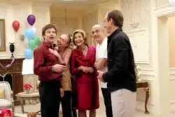 A man wearing a red shirt is embraced by his family, consisting of two women and two other men, as the majority of them stare off-screen.