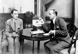 two middle-aged white men sitting on upholstered chairs next to a small, circular table; one appears worried, while the other (Grossmith) points demonstratively at a small notebook on the table.