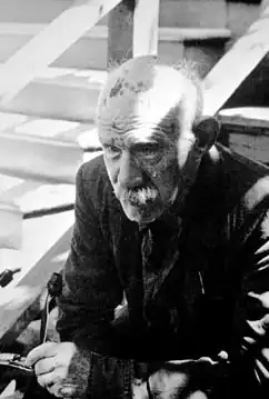 Darger in old age with a mustache, sitting down on a stairway
