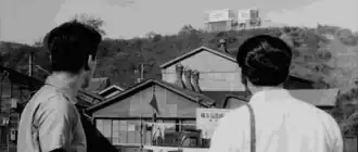 A picture showing two detectives looking up at Gondō's house on the hill, with an array of smaller rickety houses in the foreground