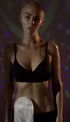 A woman with a shaved head, scars and a stoma looks at the mirror.