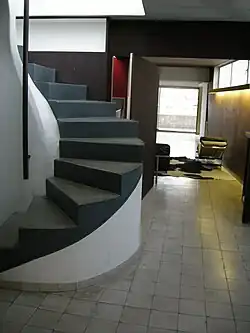 A curved concrete staircase with dark gray steps winds upward in a minimalist interior with tiled floors, wood-paneled walls, and modern furniture near a bright window