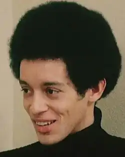 Three-quarter profile colour still from a video, of a smiling, clean-shaven young man with afro hairstyle.and black roll-neck sweater
