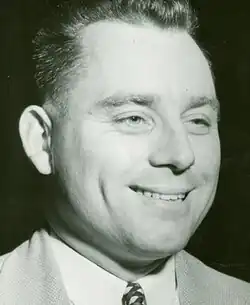Black and white photo of a middle-aged man with dark hair, wearing a white collar dress shirt, dark necktie, and a light-coloured jacket