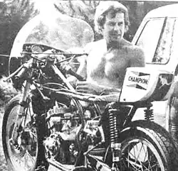 Black and white image of a man stripped to the waist in strong sunshine, alongside a part dismantled racing motorcycle