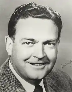 Black and white image of middle-aged man with dark hair, wearing a tweed suit jacket, white-collar shirt, and dark necktie