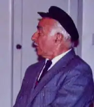 Profile of Jozo Tomasevich, a gray-haired man wearing a blue suit, a tie and a flat cap.