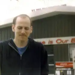 Photo of Adam Demirjian's father in front of a black gas station with a white roof. He is wearing a black hoodie which is slightly open, revealing a blue t-shirt. He appears to be walking away from the gas station. His eyes are closed.