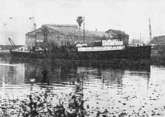 A ship underway, heading to the right. There is a crane on her stern, and two decks of superstructure above the hull. In the foreground are some shrubs; in the background is a warehouse.