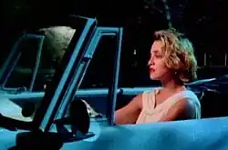 A woman with short blonde hair sits in the driver's seat of a convertible car at night, looking ahead with a serious expression.