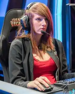 Maria Creveling playing a tournament match onstage. She is a woman with straight, long, light brown hair and brown eyes, wearing a black jacket and red undershirt.