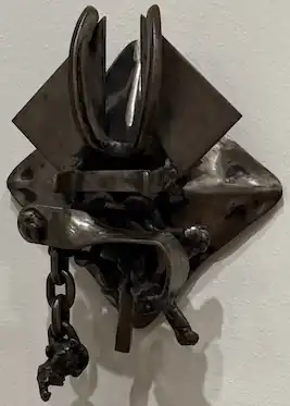 Afrophoenix No. 1, an abstract metal sculpture by Melvin Edwards. A small, dark steel relief sculpture consisting of layered, diamond-shaped pieces of metal overlaid with several other metal scraps hangs flat on the wall. Pieces of steel are bolted to the bottom portion of the work, the top is bisected by a bent horseshoe, and a short chain hangs from the lower left with lumps of metal dangling at the end.