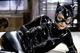 A woman reclining on a bed, wearing shiny black bodysuit with visible white stiches, and a mask including cat ears.