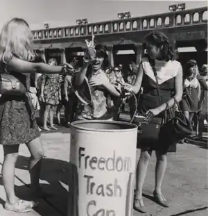 Two women toss items into the Freedom Trash Can while a female reporter looks on.