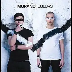 Two men sitting in front of a white wall. The one on the left is wearing a black t-shirt with a diagonal streak of white paint and the one on the right is wearing a white t-shirt with a diagonal streak of black paint.