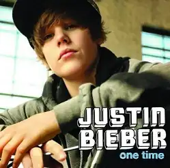 A 15-year old Justin Bieber stands in front of a building with block windows, while on the handlebars of a bicycle and wearing a hoodie and fitted hat.