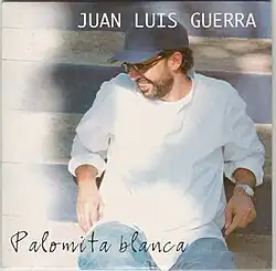 Image of Juan Luis Guerra wearing a white shirt, dark blue cap, sitting down and facing right.