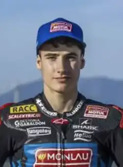 Head and shoulders view of a slim young caucasian male against a fuzzy background wearing a blue baseball cap and multi colored racing leathers with sponsors' logos