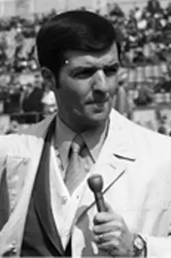Black and white photo of an adult male wearing a white-coloured suitjacket holding a microphone