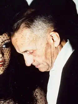 An older man with graying hair and formal attire leans toward a figure with a mannequin head with dramatic makeup