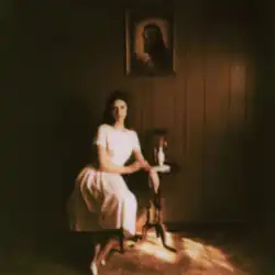 Ethel Cain sitting in a white dress beneath a picture of Jesus Christ.