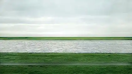 A photograph of the Lower Rhine river flowing horizontally through green fields under an overcast sky in Germany