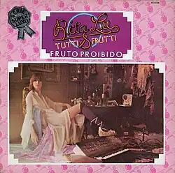 A woman sitting in a room dominated by dark colors, and wearing a white silk nightgown and stockings with a slit on the left leg, revealing a glimpse of her thigh. She is seated next to her keyboard in front of her grandmother's vanity in a boudoir setting.
