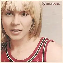 An image of a woman with a blonde bob cut and wearing a red jersey. The names of the single and the artist are superimposed over the image.