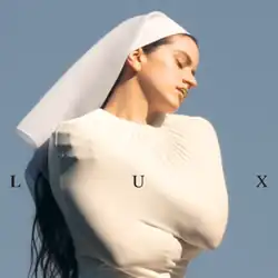 Rosalía in a white nun's habit and veil, with long dark hair, eyes closed, golden lips, hugs herself against a blue background. Word "LUX" in the centre