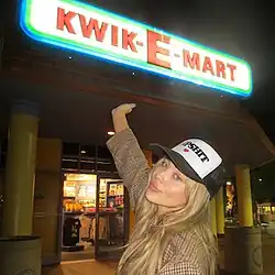 Sabrina Carpenter showing her hand to a store with a neon sign reading 'Kwik-E-Mart', wearing a cap reading 'Dipshit'.
