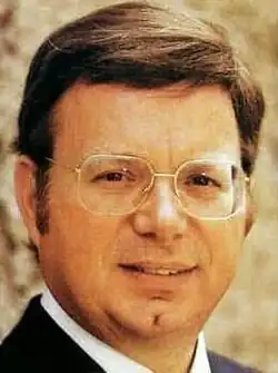 Colour portrait photo of a smooth-shaven, half-smiling man with short brown hair, spectacles and sideburns.