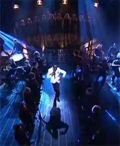Solo singer in white coat surrounded by backing choir, bright spotlights above