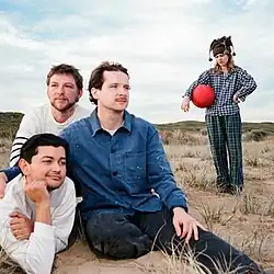 At a beach, three men sit on the sand, contentedly dazing in different directions. In the background, a woman holding a red dodgeball stares at the ground, looking disappointed.