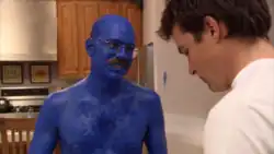 A mustached man painted in blue smiles at another man, whose back is faced towards the camera