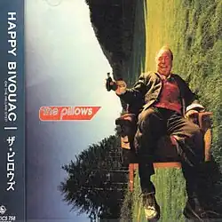 A happy man sits in an overturned chair in a green field holding a liquor flask.