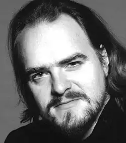 Black and white studio portrait of a man with high forehead, long dark hair, and trimmed beard and moustache. He is looking intently at the camera.