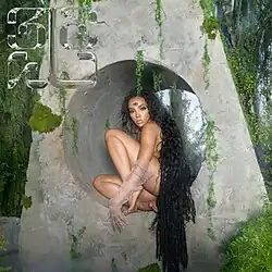 Tinashe sitting nude in a kneeling position within a moss-covered gray stone tunnel, with long dark hair and wearing a sheer glove on her left arm, set against a backdrop of lush green foliage