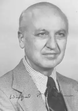 A balding man in a checkered suit with a striped shirt and dark tie