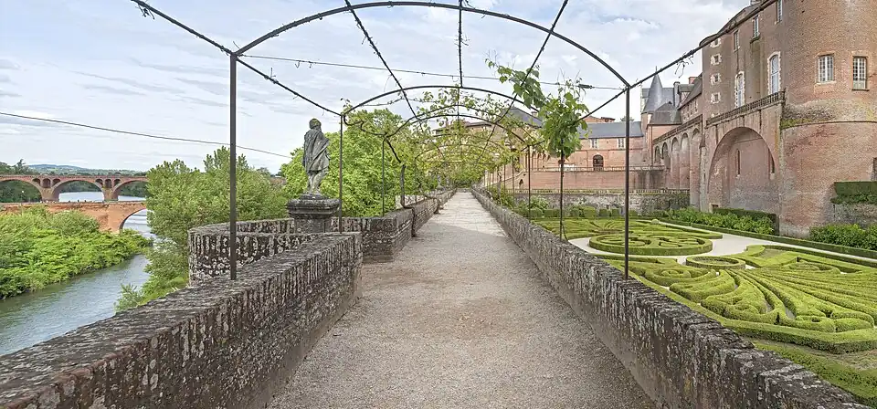 Photo couleur d'une allée sablée ombragée d'une tonnelle de fer forgé recouverte de vigne. La promenade marque la limite entre un jardin en contrebas à droite et le vide des anciens remparts à gauche.