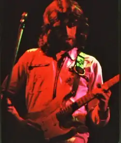 Portrait en buste d'un homme barbu et chevelu jouant d'une guitare au cours d'un concert.