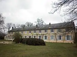 Photographie du bâtiment principal de la Colonie, à Condé-sur-Vesgre. Il s'agit d'un long bâtiment symétrique à un étage, avec des grandes fenêtres régulières, six de chaque côté de la porte. Devant, un petit terrain pelousé avec des buissons et quelques arbres.