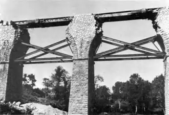 Photographie en noir et blanc du viaduc de profil montrant le tablier installé entre les deux arches détruites avec les piles consolidées par des poutres montées en croix de Saint André.