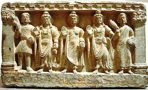 Une triade précoce du bouddhisme mahāyāna. De g. à d. un dévot kouchan, le Bodhisattva Maitreya, le Bouddha, le Bodhisattva Avalokiteshvara, et un moine bouddhiste. IIe-IIIe&nbsp;siècle, Gandhara.