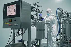 Personne portant une combinaison blanche de salle blanche, des gants bleus et un masque, utilisant un équipement mobile en acier inoxydable pour le nettoyage d’un bioréacteur dans un environnement stérile