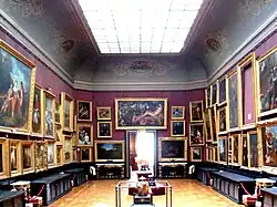 Grande salle aux tentures rouges couvertes de tableaux de grande dimension avec une porte d'entrée au fond